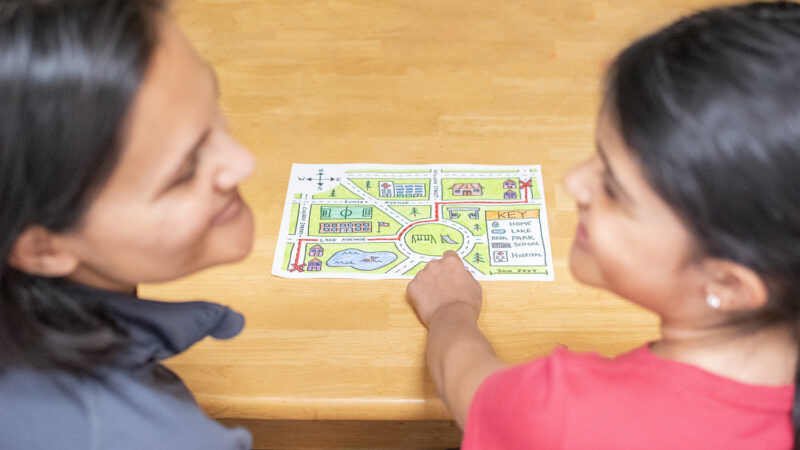 A mother and child sit at a table together, talking. The child is pointing to a small map they made together.