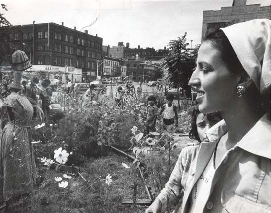 A woman in the foreground of an urban community garden is seen, profile, smiling. In the background is a group of working community gardeners,