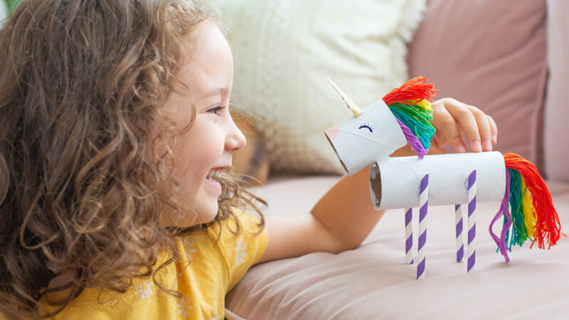 A young child holds a small unicorn she made from paper tubes and other craft supplies.