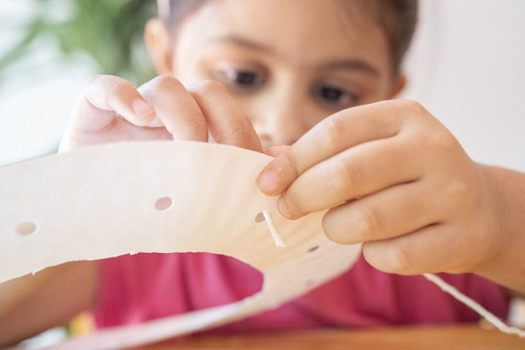 A young child threads yarn through holes punched in a paper plate. 