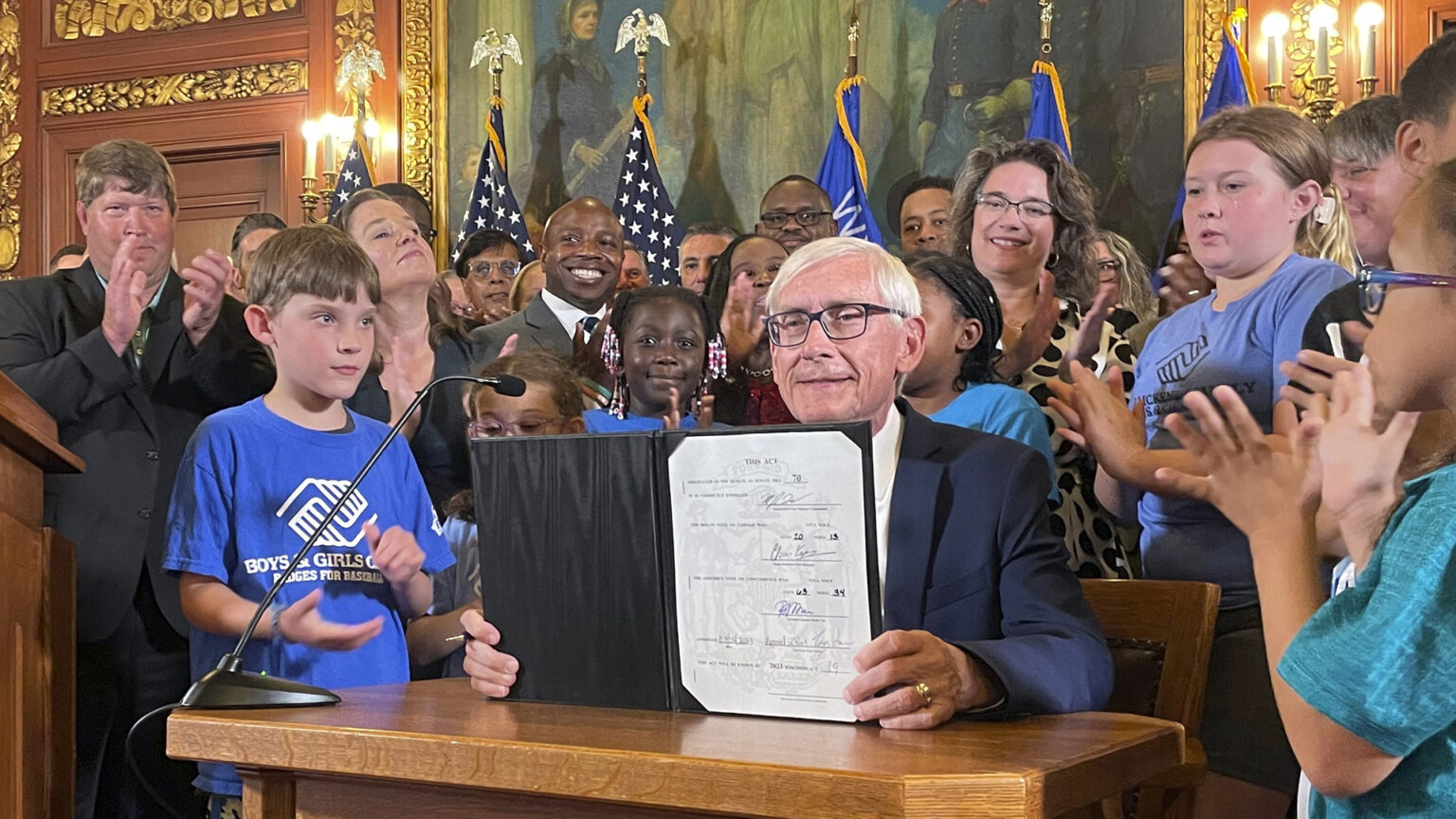 Tony Evers sits at a small wood table and displays a series of signatures in a document inside an open folder, with children and adults applauding while standing beside and behind him in a room with a row of U.S. and Wisconsin flags, electric wall sconces, paintings, and filigree gilded wall decorations.