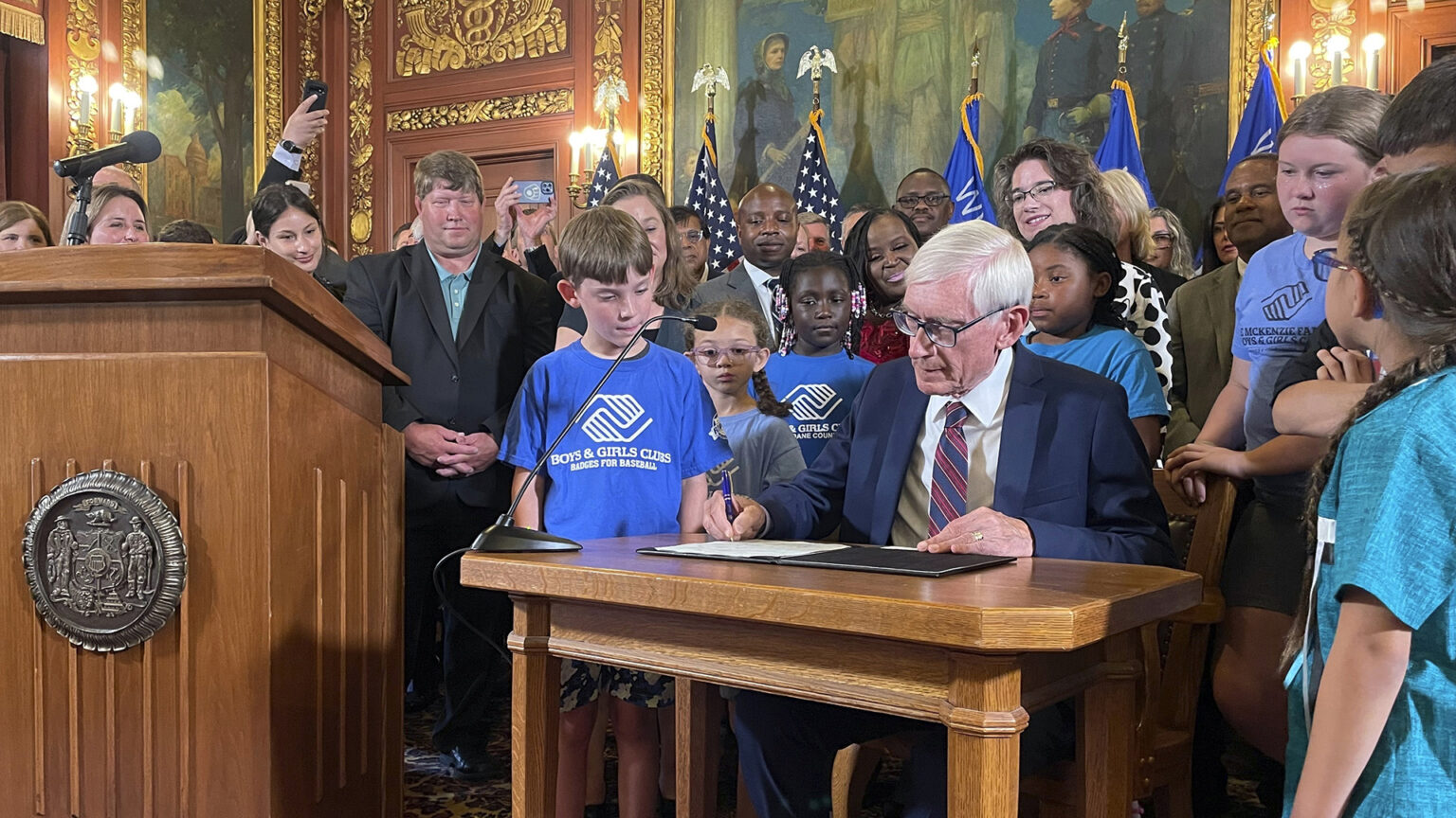 Tony Evers sits at a small wood table placed next to a wood podium affixed with the Great Seal of the State of Wisconsin and uses a pen to sign a document in a folder, with children and adults standing beside and behind him in a room with a row of U.S. and Wisconsin flags, electric wall sconces, paintings, and filigree gilded wall decorations.