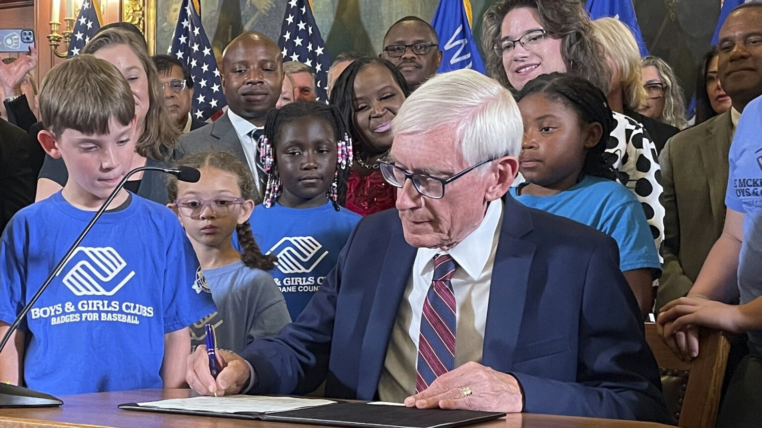 Tony Evers sits at a small wood table and uses a pen to sign a document in a folder, with children and adults standing beside and behind him in a room with a row of U.S. and Wisconsin flags, electric wall sconces, paintings, and filigree gilded wall decorations.