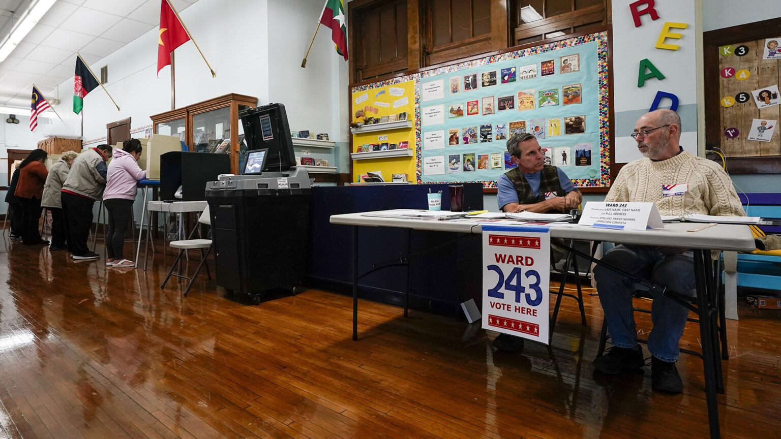 Two people sit at a folding table with a sign reading Ward 243 and Vote Here affixed to its front, with other people standing and filling out ballots in privacy booths next to ballot tabulator machine in a room with polished hardwood floors, glass-doored cabinets, flags of different nations mounted on its walls, and bulletin boards showing a variety of graphics.