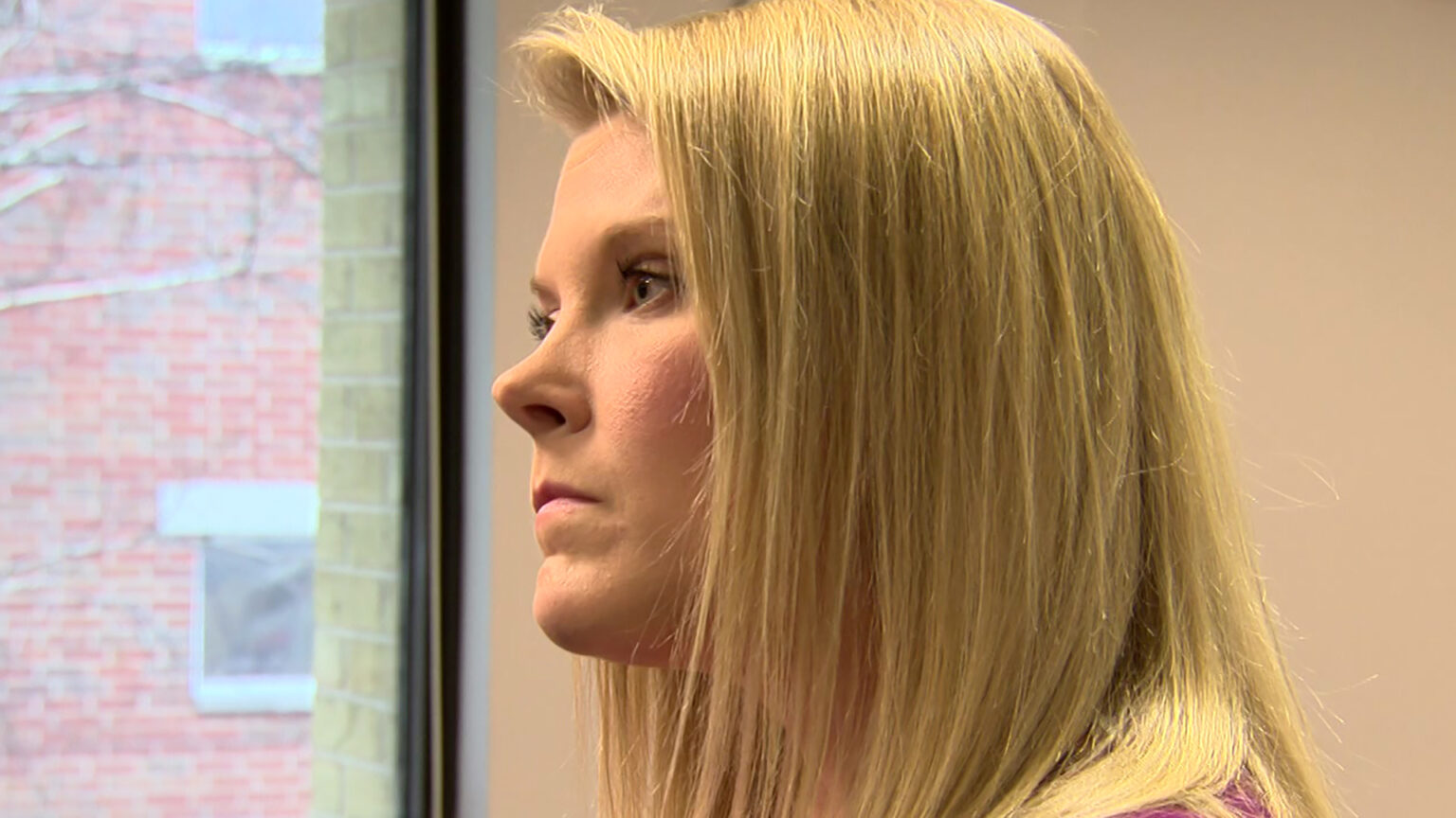 Meagan Wolfe stands and listens in a room with a window showing tree branches and the exterior of an adjacent brick building.