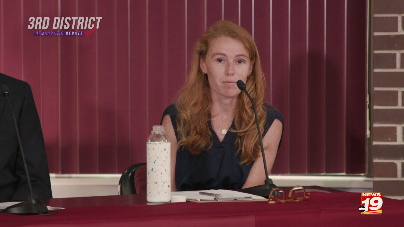 Rebecca Cooke speaks into a microphone while sitting at a table, with a water bottle, clipboard and glasses in front of her, in a room with a brick wall and closed vertical blinds in a window, with graphics on the image reading 3rd District Democratic Debate and ABC News 19.