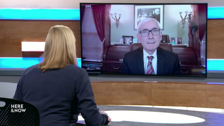 Frederica Freyberg sits at a desk on the Here & Now set and faces a video monitor showing an image of Tony Evers.