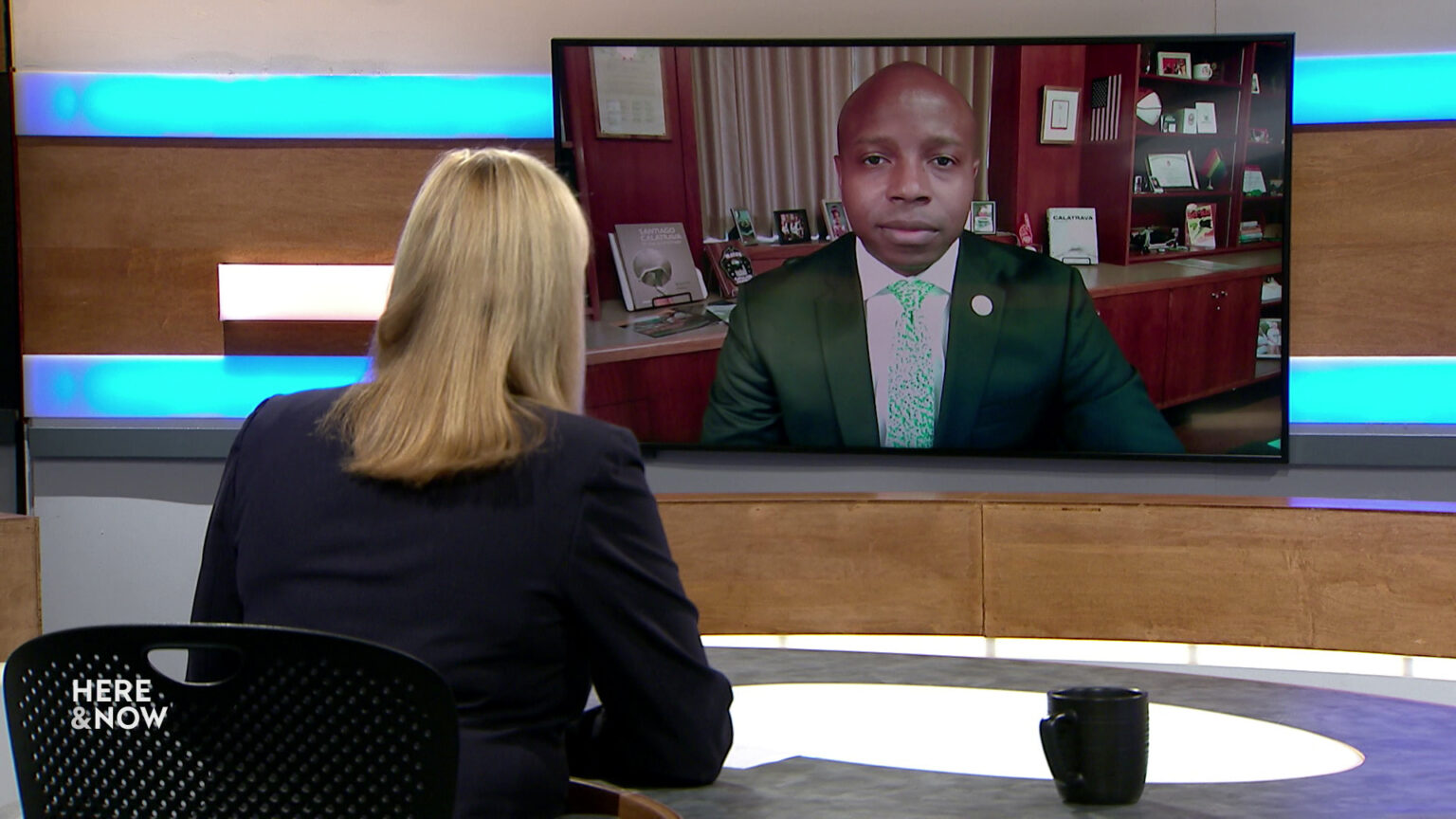 Frederica Freyberg sits at a desk on the Here & Now set and faces a video monitor showing an image of Cavalier Johnson.