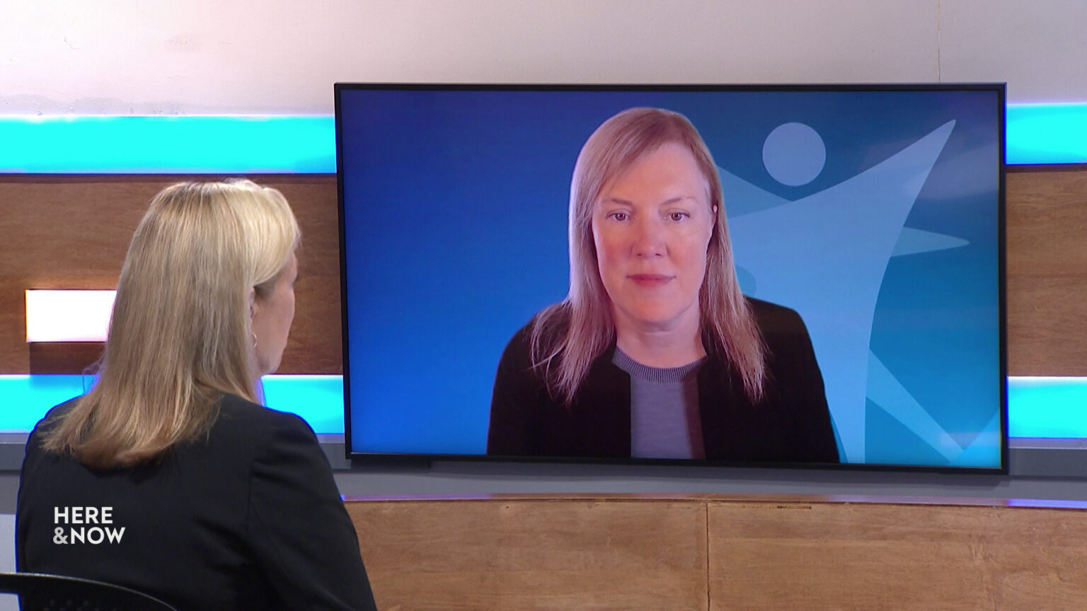 Frederica Freyberg sits at a desk on the Here & Now set and faces a video monitor showing an image of Kirsten Johnson.