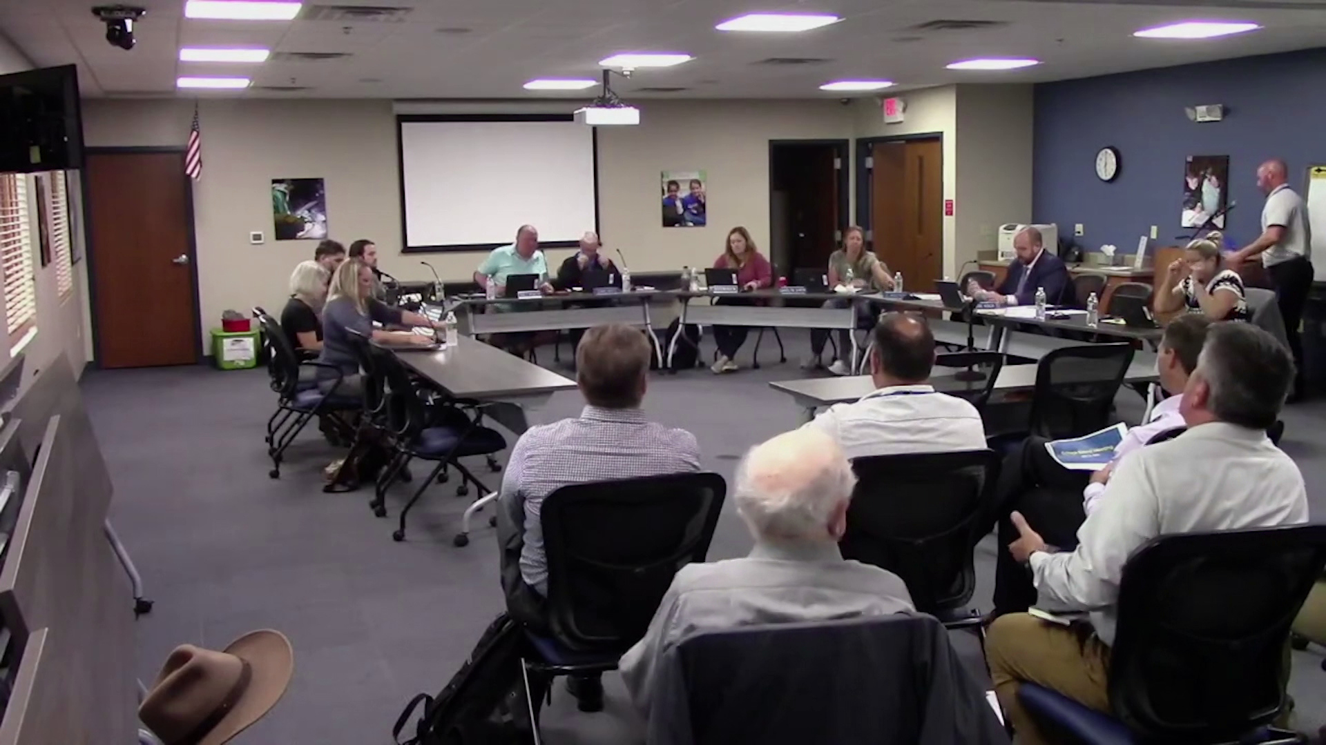 Members of the Mukwonago school board sit in rolling chairs around multiple folding tables arranged in a horseshoe position, with other people sitting and facing the meeting, in a room with a projector screen, exit sign and fluorescent lights.