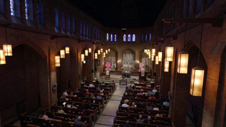 An overhead view in the interior of a church shows people sitting in rows of wood pews facing an altar with a stepped platform, two pulpits and altarpieces with crosses, in a nave with brick-lined pillars framing gothic arches with mounted hanging lamps, and supporting a line of stained-glass windows surrounding the space.