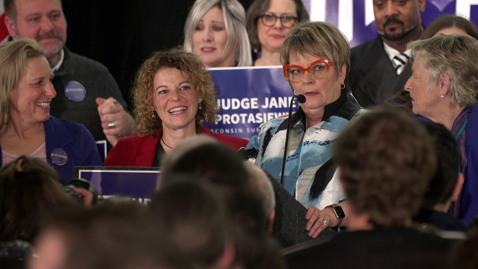Janet Protasiewicz speaks into a microphone affixed to a podium while Jill Karofsky, Rebecca Dallet and Ann Walsh Bradley stand on either side of her, surrounded by other people behind them and facing them.