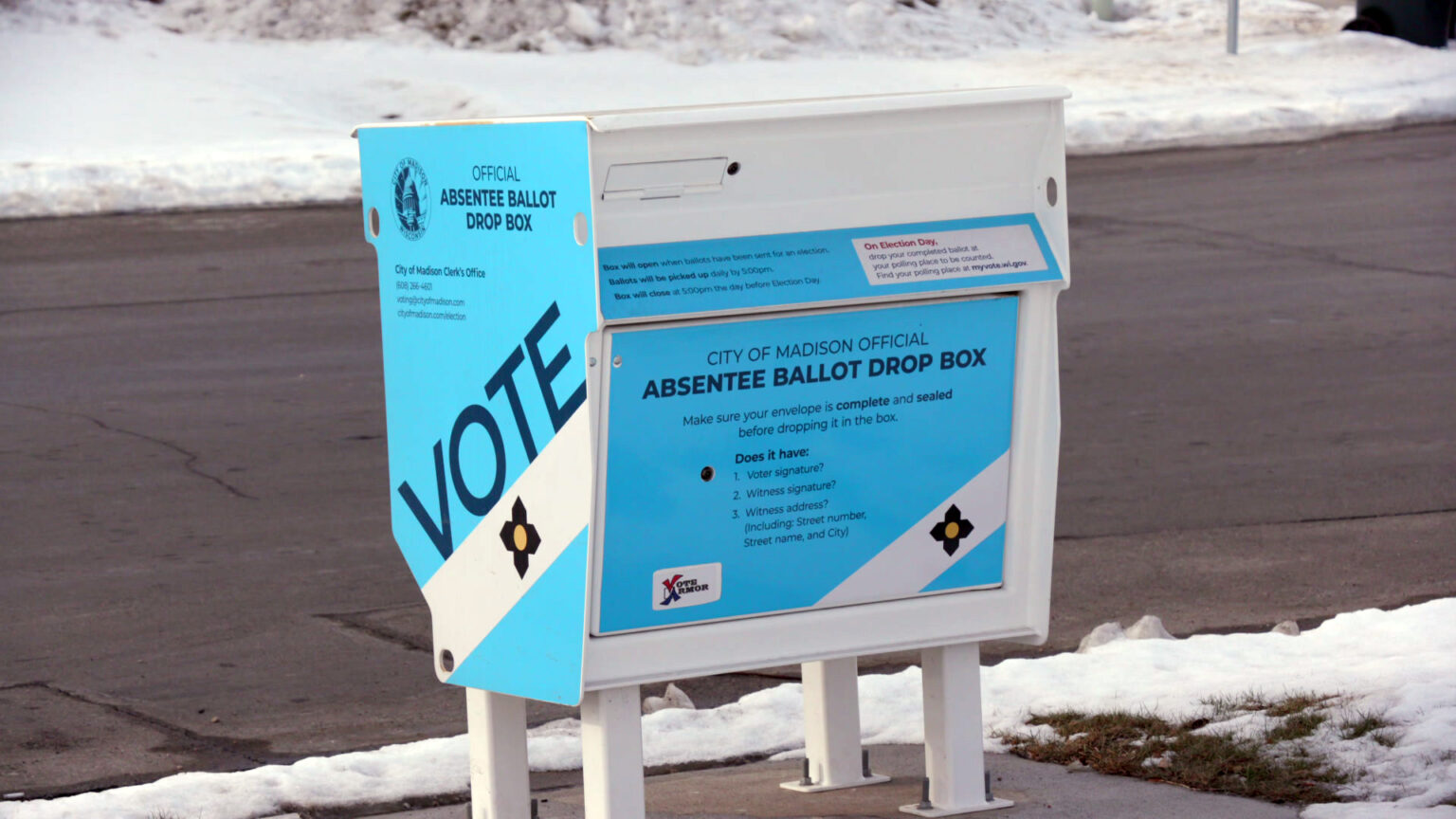 An absentee ballot drop box bolted to concrete next to a street stands amid melting snowdrifts.