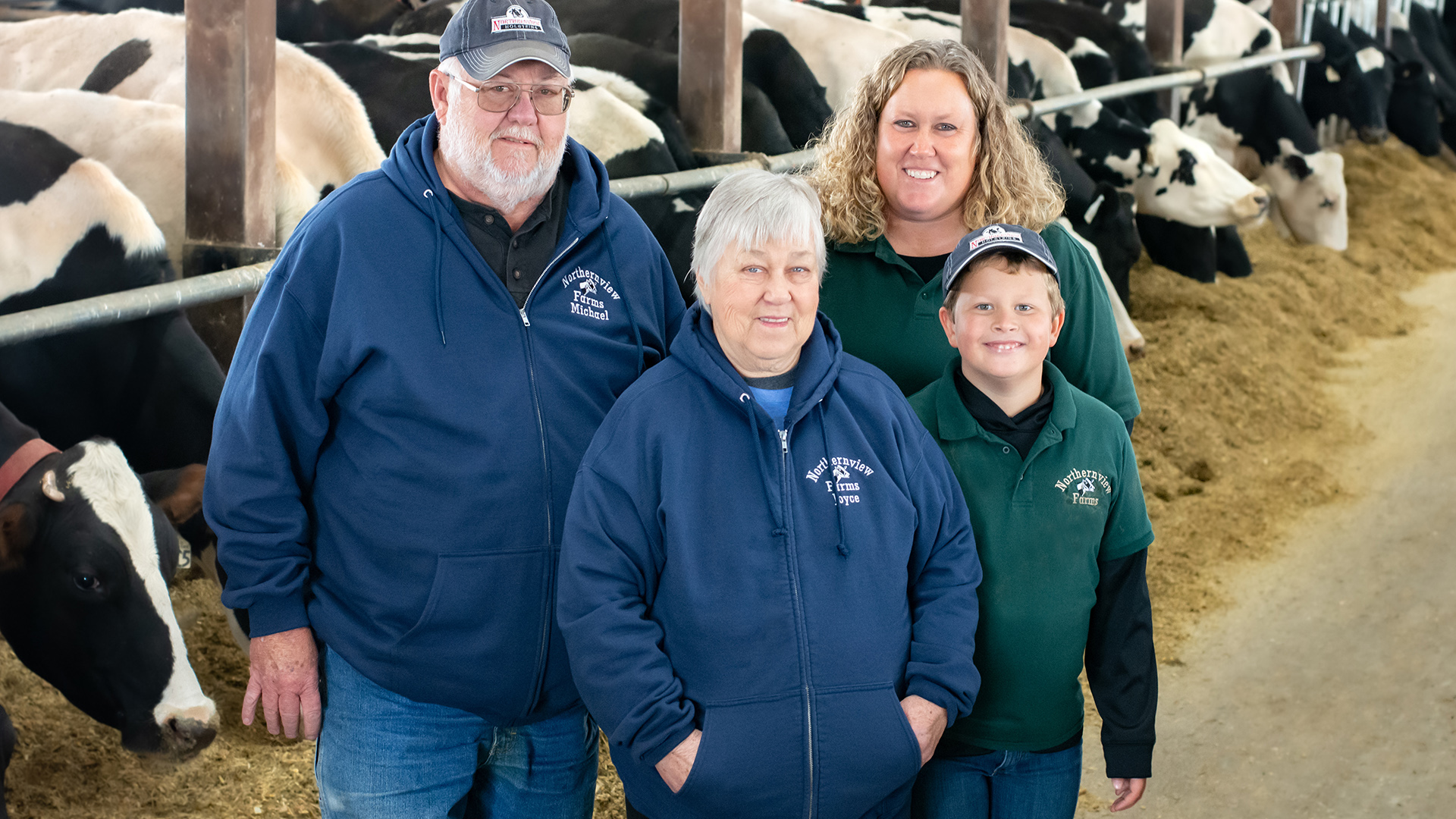 Michael, Joyce, Sara and Noah Byl in a cattle barn with multiple Holstein cows standing at a rail and eating feed.