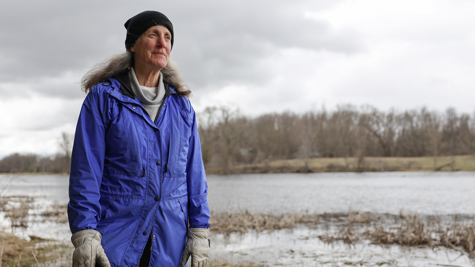 Lisa Doerr stands outside in front of a small body of water, with wetlands in the foreground and a tree-lined shore in the background under an overcast sky.