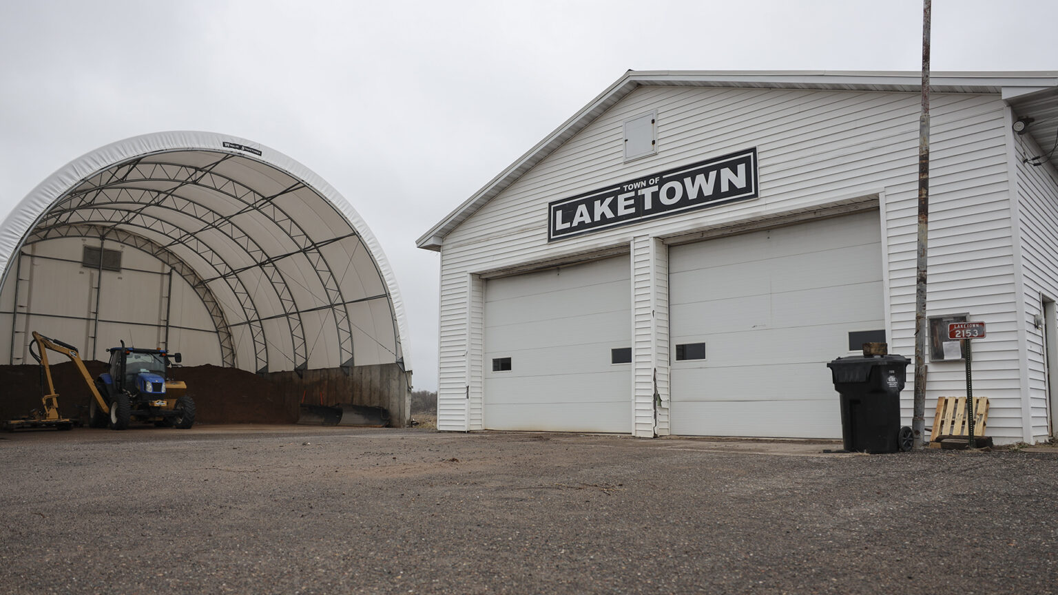 A building with tall double garage doors and a sign reading Town of Laketown stands next to a half-round shed with metal framing and a plastic roof and rear wall.