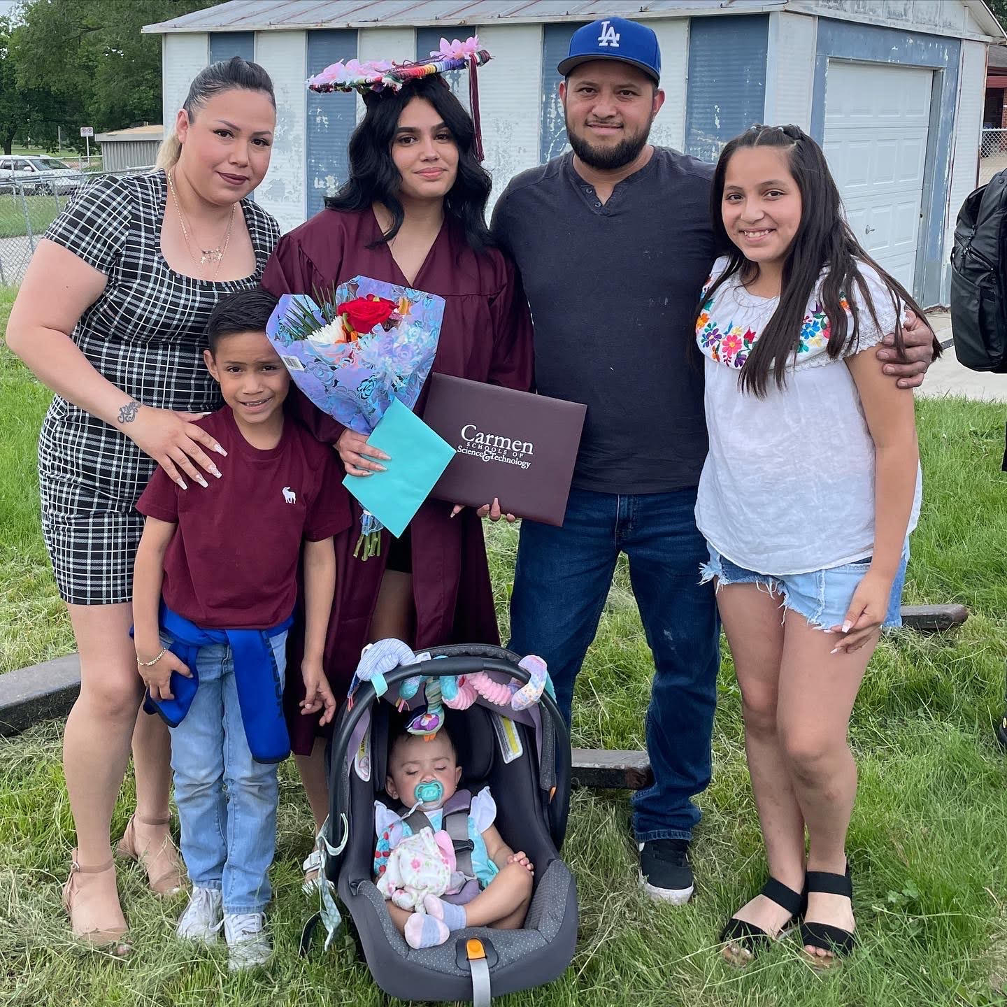 Diana Rico poses for a photo alongside her family while standing in a grassy field, with a garage, fencing and trees in the background.
