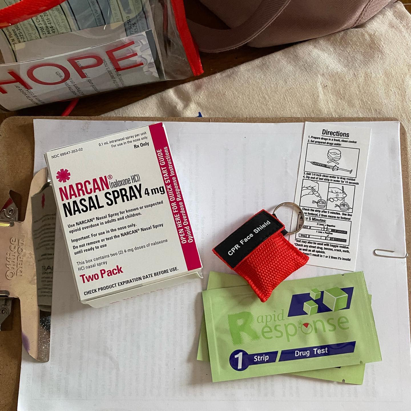 A box of Narcan nasal spray, two Rapid Response drug tests, a CPR face shield and instruction manual sit on a clipboard with a piece of paper, next to a transparent plastic pouch with the word "HOPE" on its side.