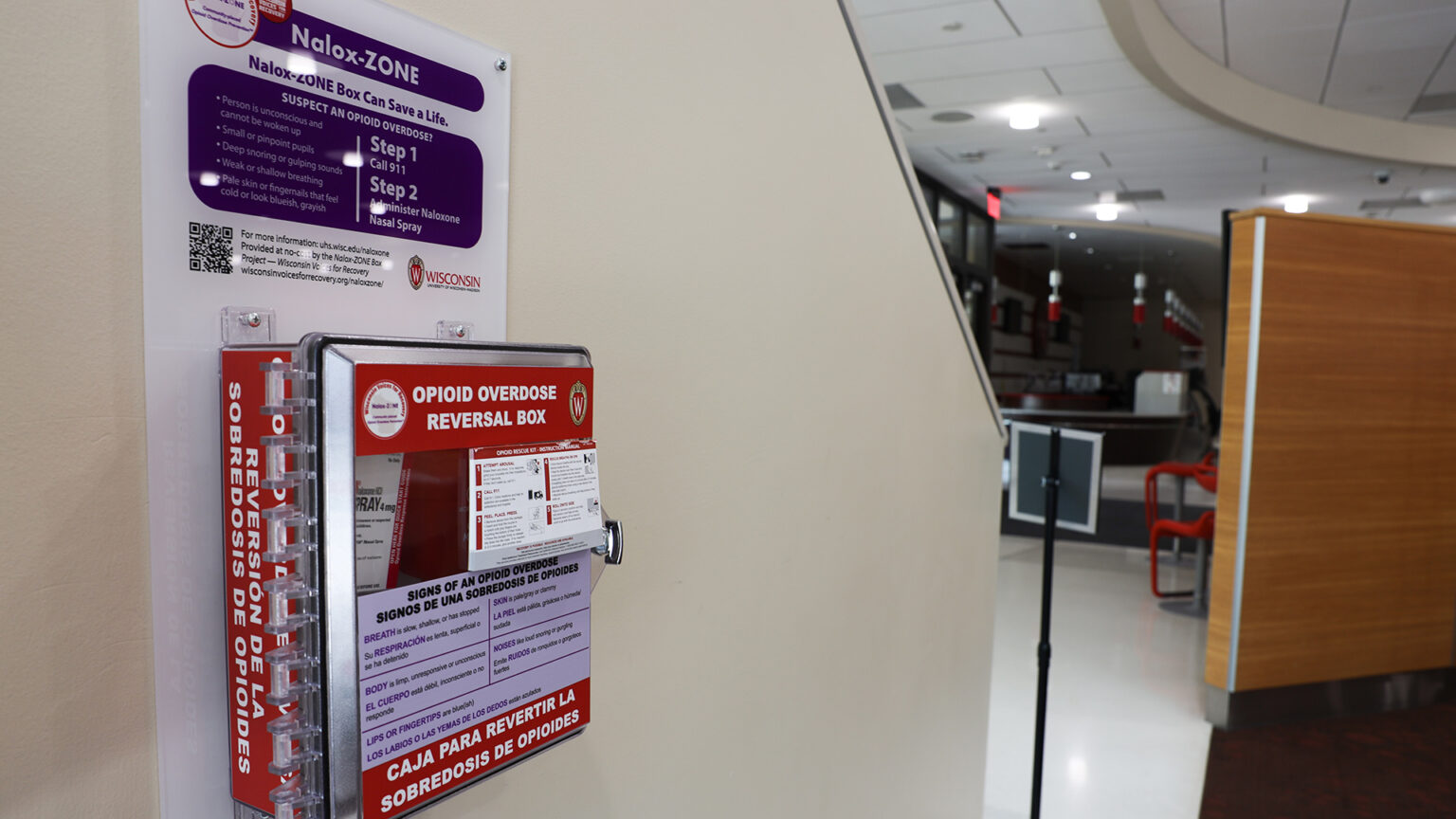 A wall mounted metal-sided box with the label Opioid Overdose Reversal Box and instructions in text of different sizes is mounted on a wall alongside a plastic sign with the label Nalox-ZONE in a room with food service counters in the background.