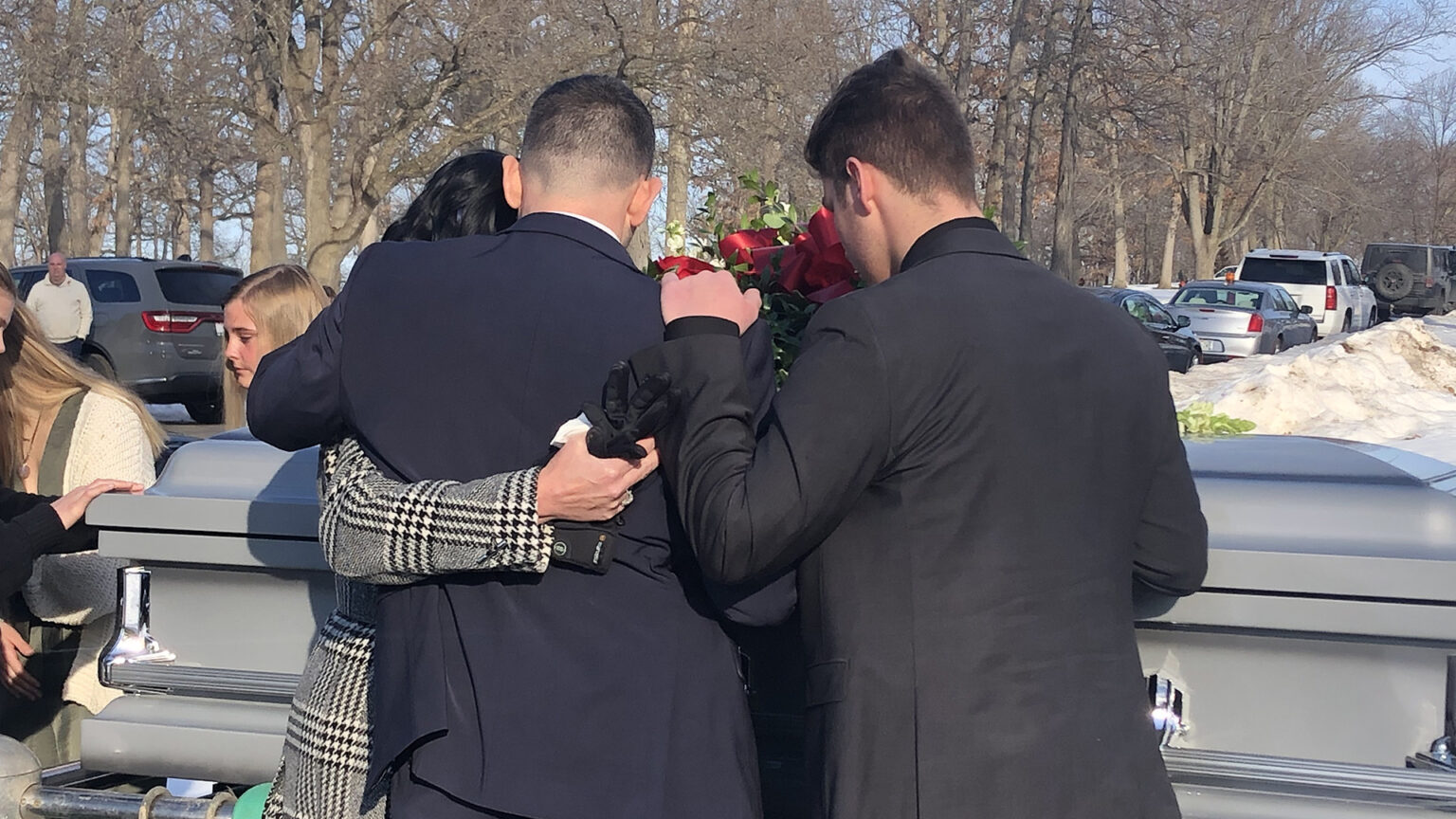 Erin, Rich and Caden Rachwal hold their arms around each other and bow their heads while facing a closed casket while standing outdoors, with other people, parked cars, leafless trees and snow drifts in the background.