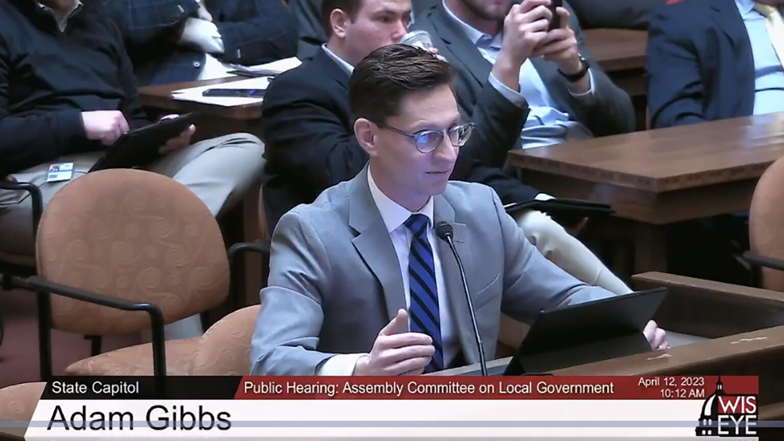 A screenshot shows Adam Gibbs seated at a desk while speaking into a microphone and looking at a laptop computer screen, with other people seated in the background, with a video graphic on the bottom of the image noting his name, the date and setting as a public hearing of the Assembly Committee on Local Government.