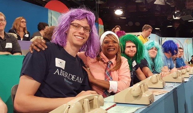 A line of smiling volunteers wearing colorful wigs and outfits sitting in front of phones in a recording studio.