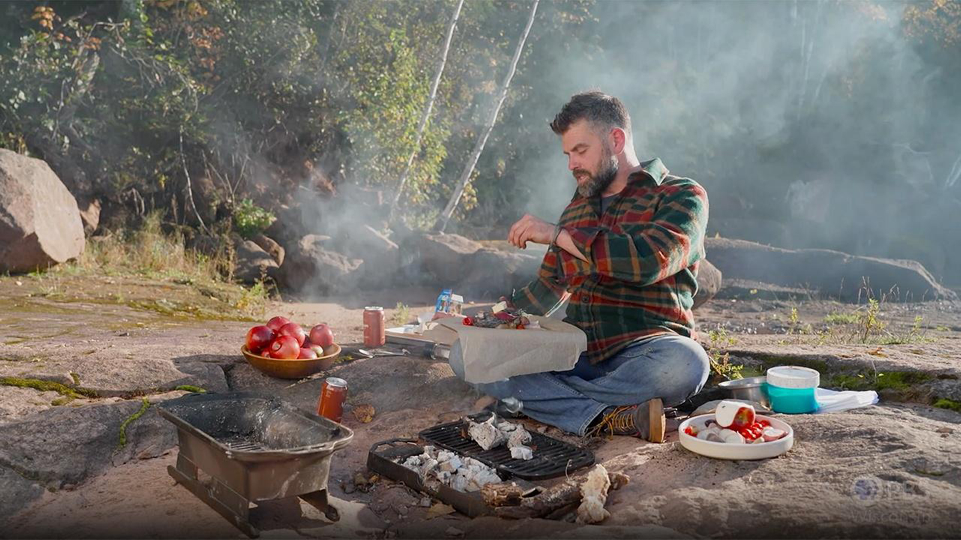 A man sits near an outdoor fire ring cooking fish and apples.
