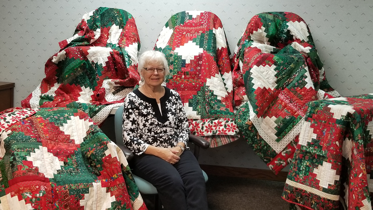 Klaudeen sits in a chair surrounded by red, white and green Christmas quilts.
