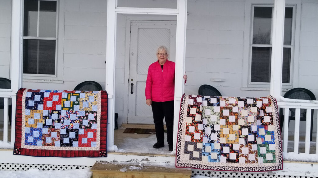 Klaudeen Hansen standing on a snowy, covered porch with two colorful patchwork quilts.