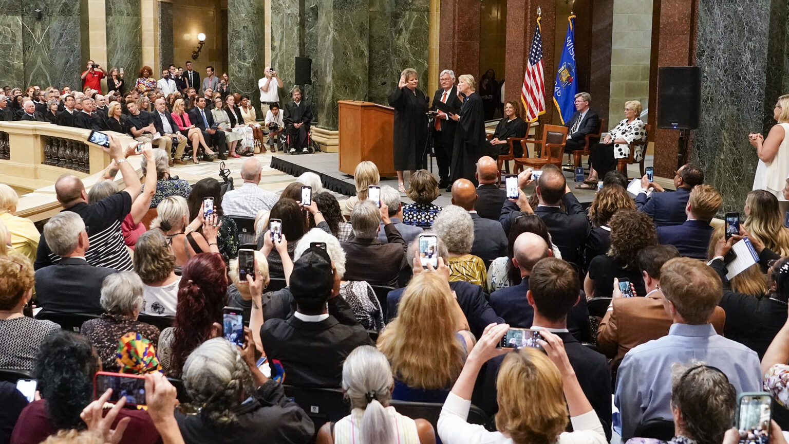 Janet Protasiewicz raises her right hand while standing next to a wood podium and facing Ann Walsh Bradley, in a circular space with marble masonry, the U.S. and Wisconsin flags and portable loudspeakers, with people seated on either side watching the proceedings and many recording or taking photos with their mobile devices.