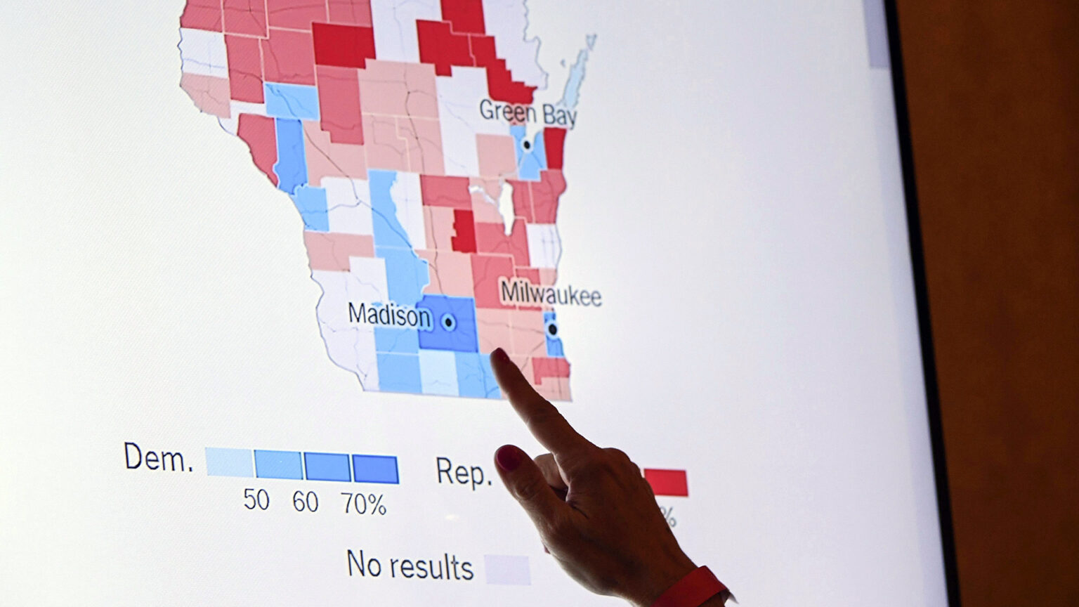 The index finger of a right hand points toward a screen featuring a map of Wisconsin showing the county-level results by percentage for Democratic and Republican candidates.