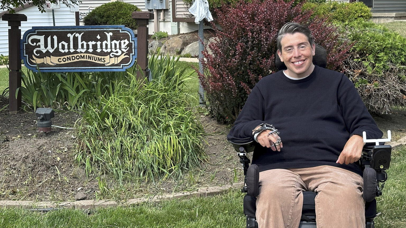 Jimmy Anderson poses for a photo outside in his wheelchair in front of a building with a wood sign reading Walbridge Condominiums placed among shrubs and other landscaping plants.