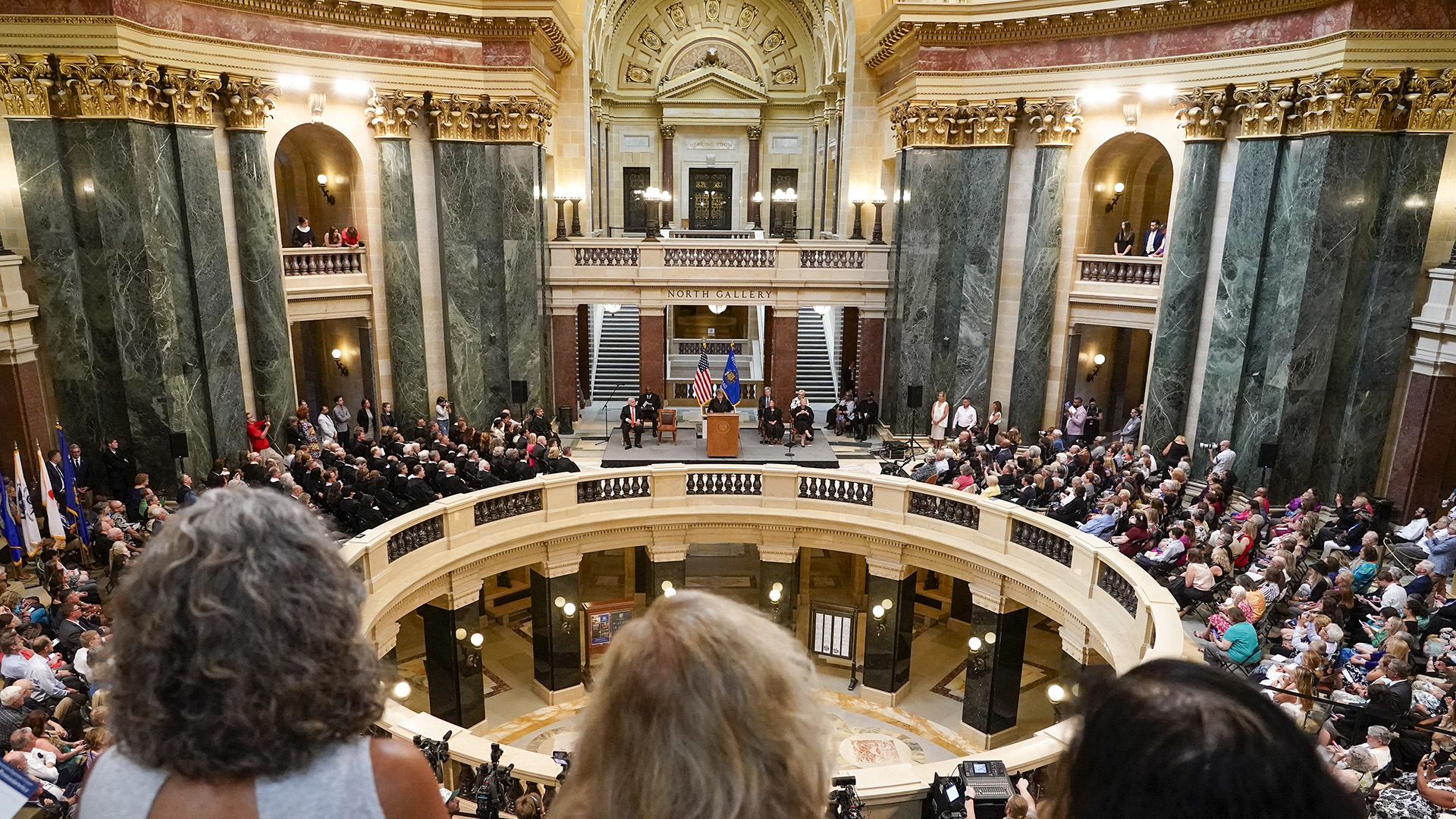 Numerous people sit and stand in the round on multiple levels of a rotunda with different colors of marble masonry, facing Janet Protasiewicz as she speaks while standing behind a wood podium and in front of the U.S. and Wisconsin flags