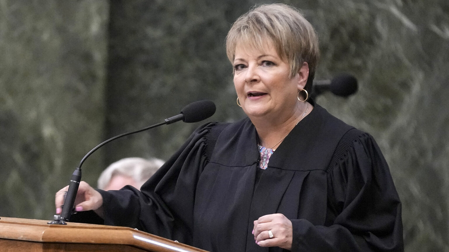 Janet Protasiewicz wears judicial robes and speaks into a microphone mounted to a wooden podium, with out-of-focus marble walls in the background.