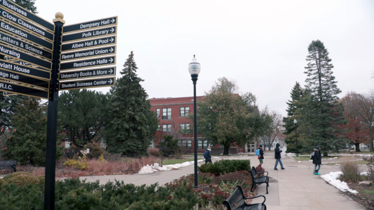 A pole affixed with more than a dozen signs bearing the names of buildings and directional arrows over looks an outdoor space with sidewalks, street lights, park benches and other landscape features, with trees and buildings in the background.