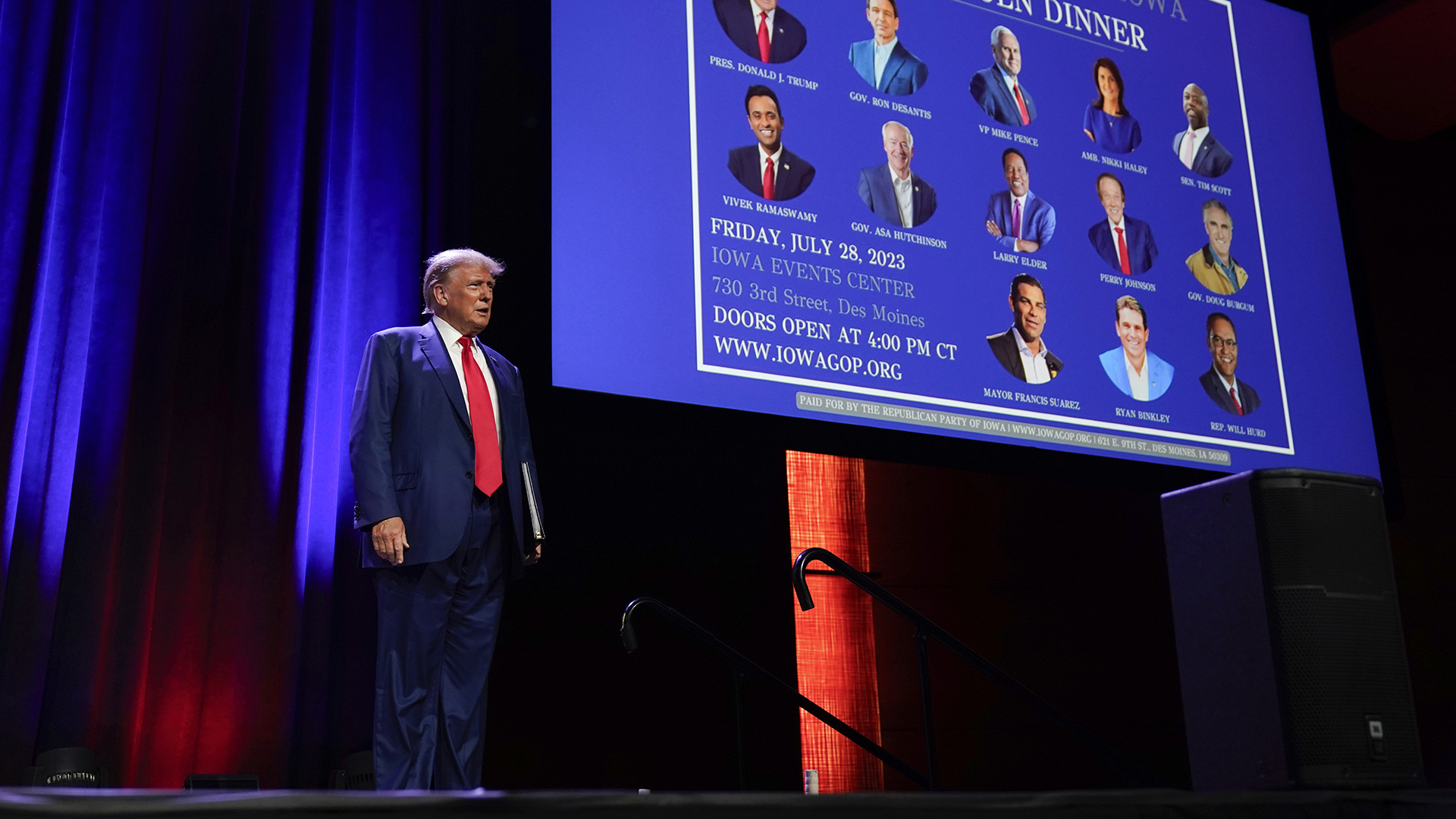 Donald Trump stands on a stage with a projector screen showing 13 candidates running in the 2024 Republican presidential primary, with a stage curtain in the background.