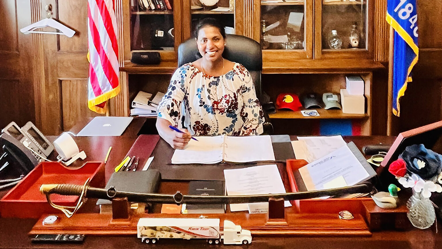 Rejani Raveendran poses for a photo while sitting in a high-backed leather chair at a wooden desk with multiple items on its surface, including a phone, speakers, various papers and books, photo frame, ceremonial sword and model semi truck, in a room with glass-doored cabinets and the U.S. and Wisconsin flags in the background.