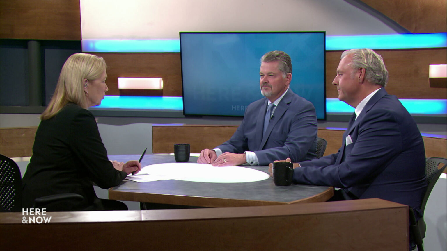 Bill McCoshen and Scot Ross sit next to each other and across from Frederica Freyberg at a desk on the Here & Now set.