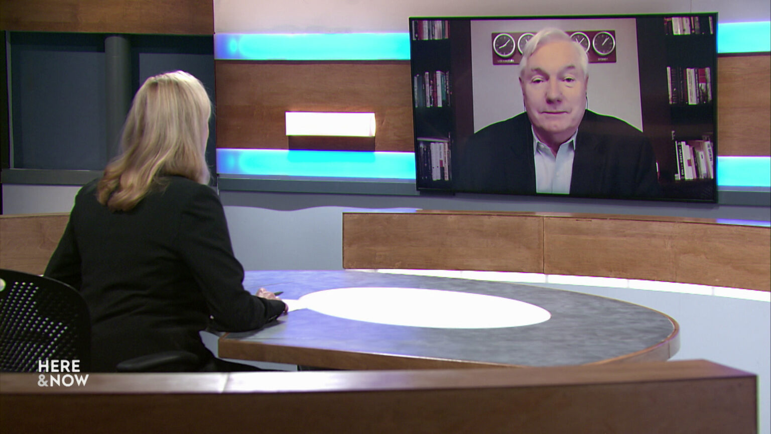 Frederica Freyberg sits at a desk on the Here & Now set and faces a video monitor showing an image of Michael Osterholm.