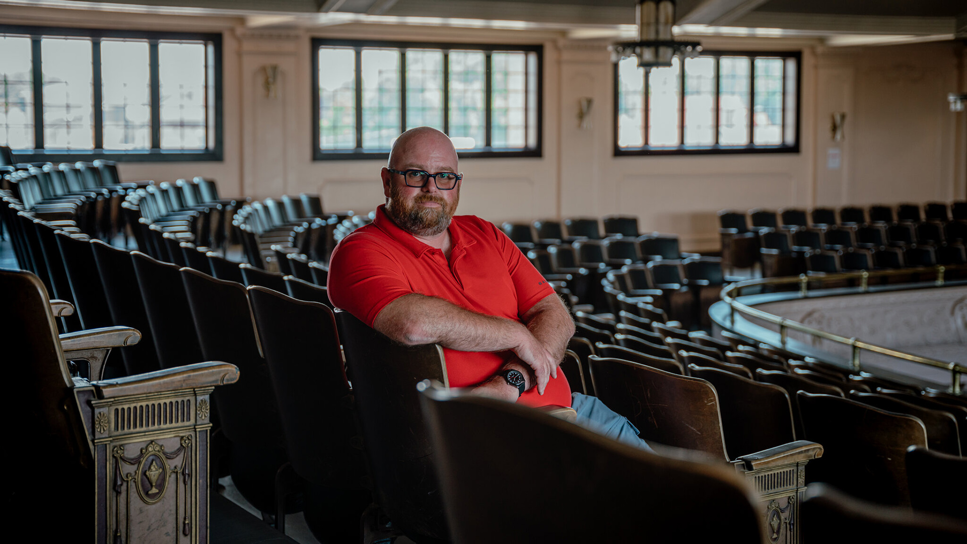Rohn Bishop poses for a portrait while seated in a wood folding chair at the end of a row of chairs in a curved balcony seating section of a multi-story room, with windows in the background.