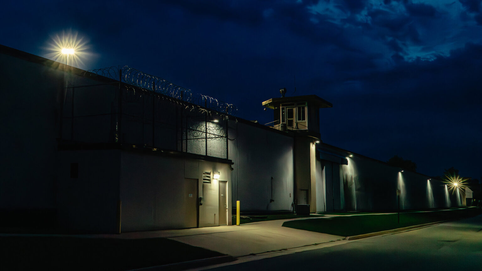 Exterior lighting illuminates the walls, towers and razor wire of a prison under a cloudy nighttime sky.