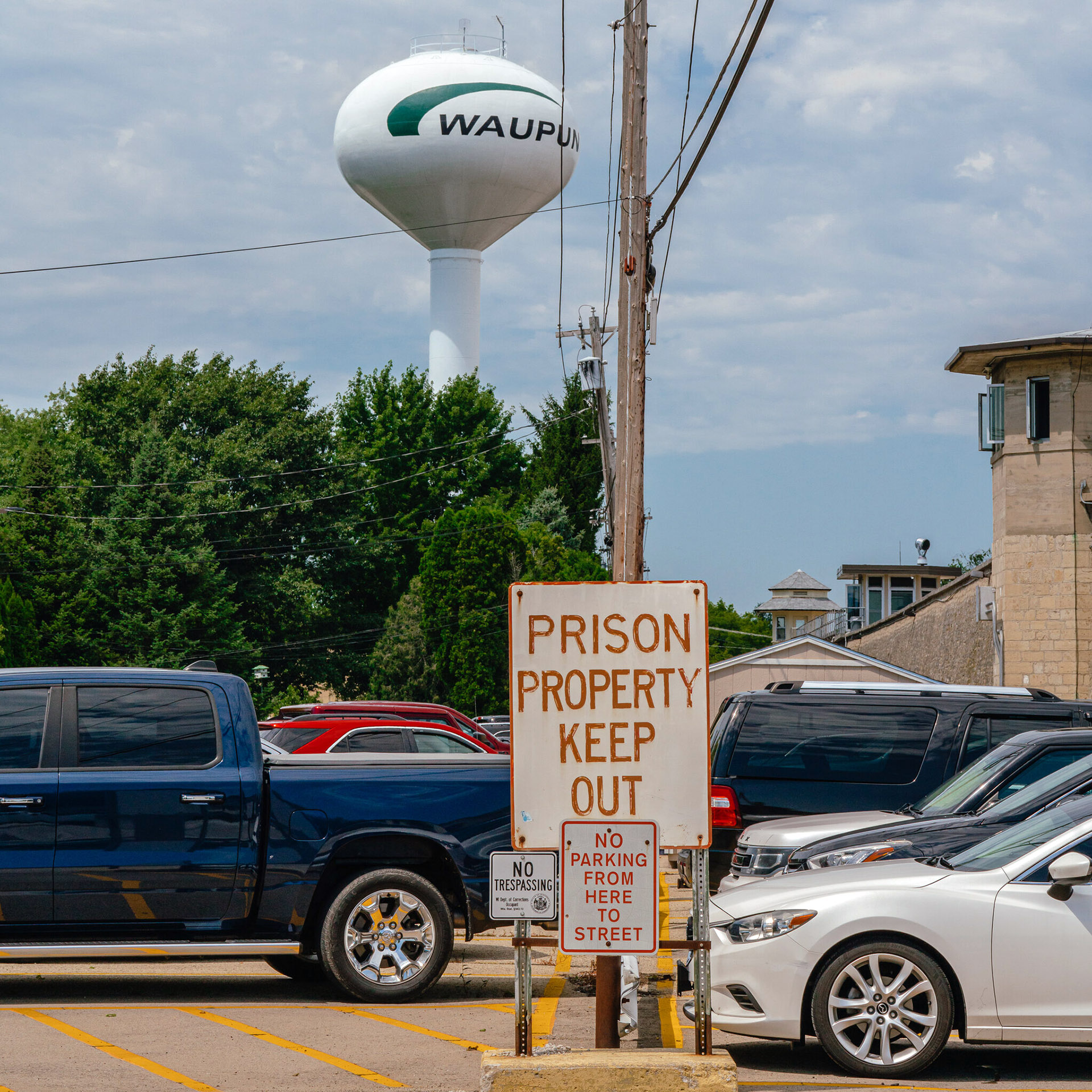 A sign reading "Prison Property Keep Out" stands in parking lot amid parked vehicles, with a utility poll, trees, building and water tower with a painted label reading "Waupun" in the background.