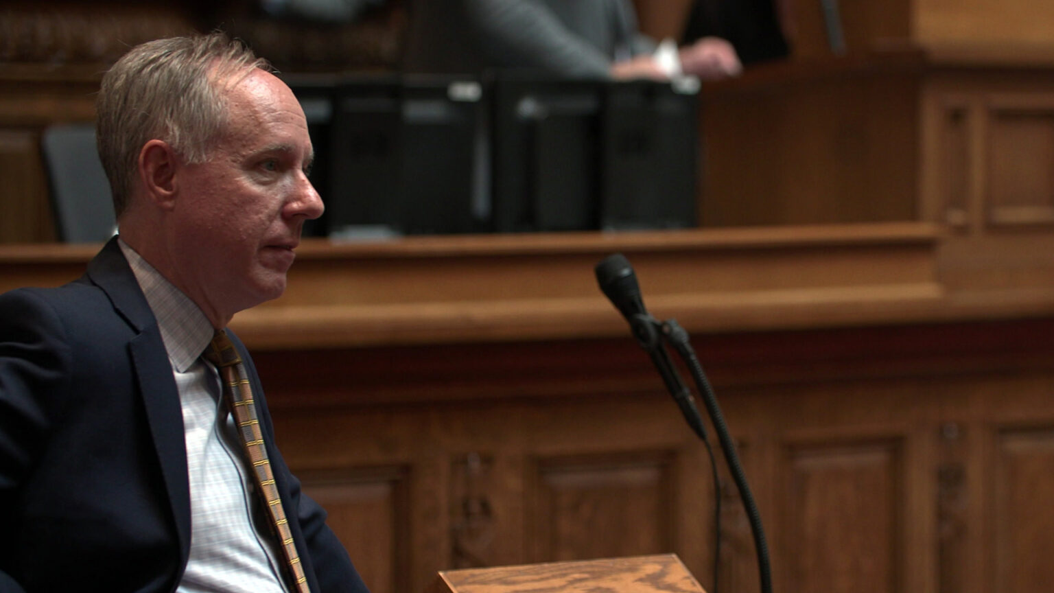 Robin Vos stands behind a wood podium with a microphone and listens, with other people standing at a legislative dais in the background.