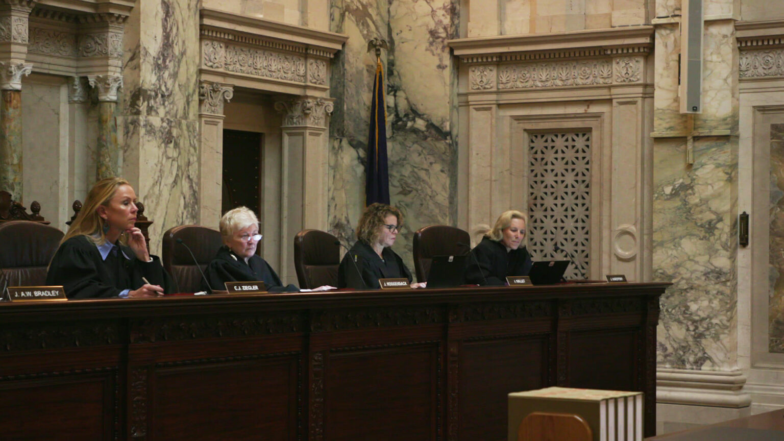 Annette Ziegler sits in her chair at the Wisconsin Supreme Court bench and holds her left hand to her face while writing with her right hand, with then-Justice Patience Roggensack along with justices Rebecca Dallet and Jill Karofsky seated to the side, in a room with marble pillars, lintels and other masonry.