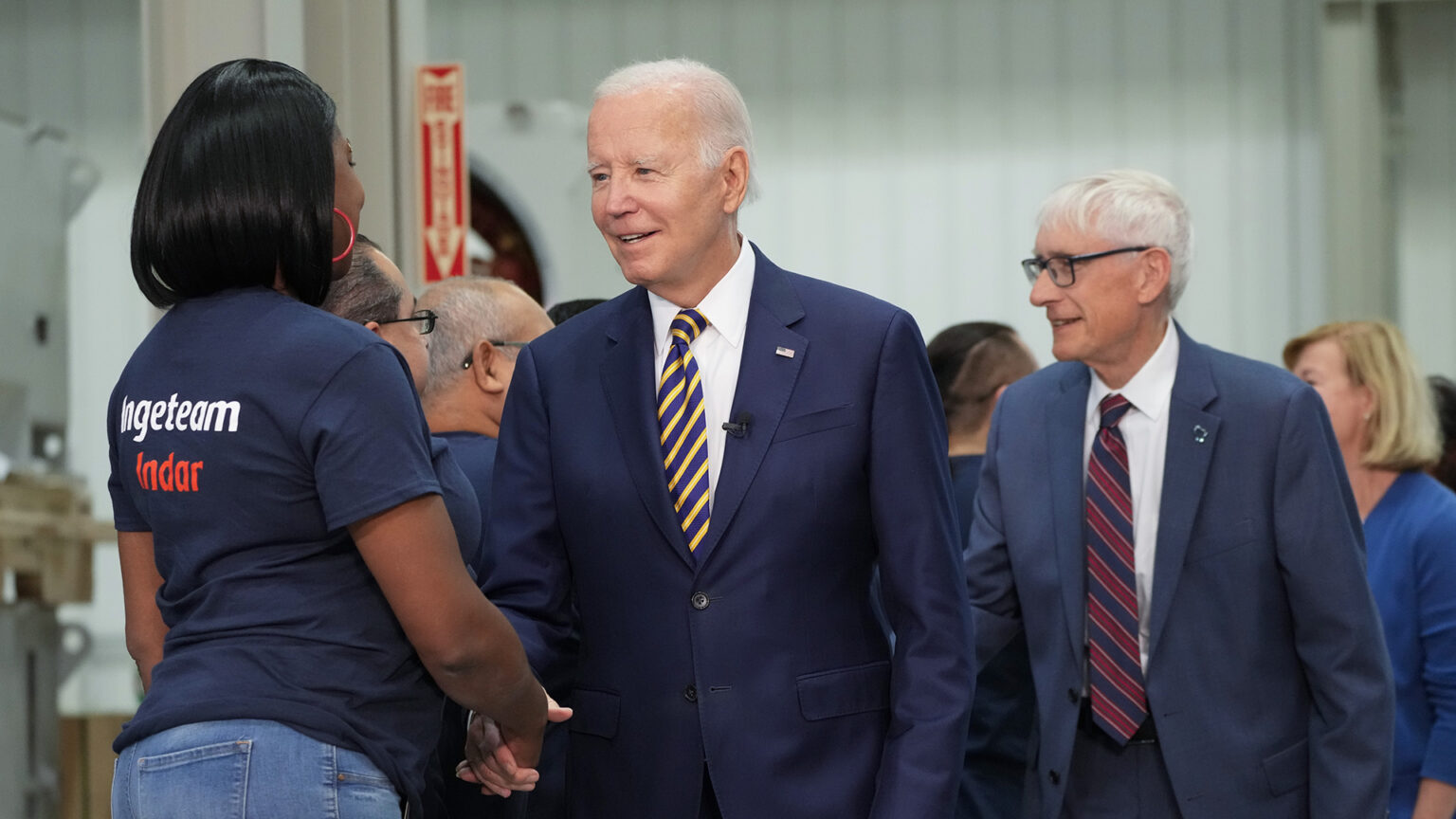 Joe Biden and Tony Evers share the hands of a line of people facing them, in a room with corrugated metal walls and industrial equipment.