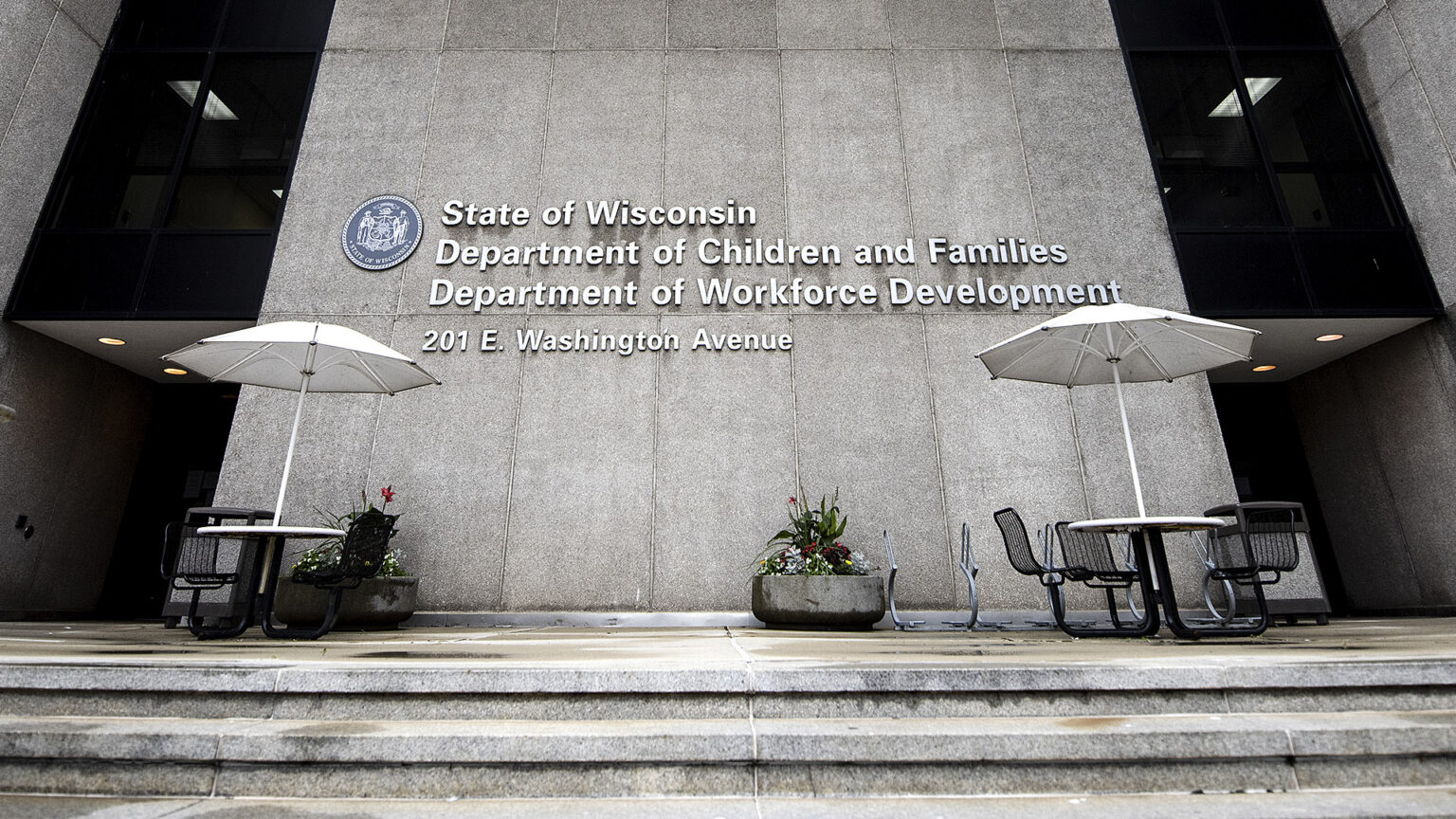 Two outdoor metal tables with attached chairs and umbrellas stand on the concrete patio in front of a building with poured concrete masonry, with the Wisconsin state seal affixed to the wall next to a letter sign reading State of Wisconsin, Department of Children and Families and Department of Workforce Development.