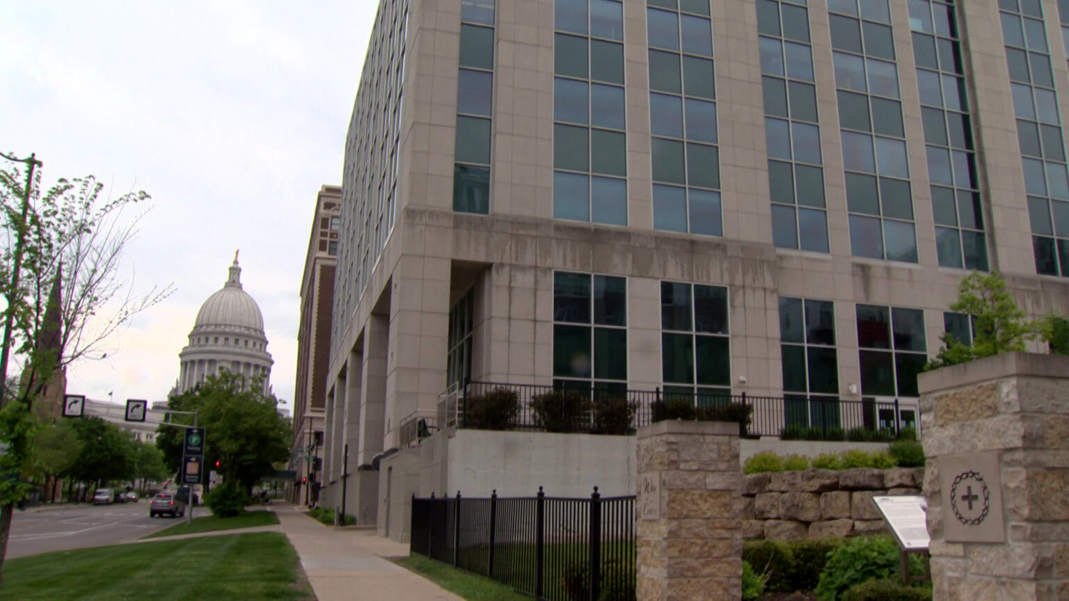 A masonry and glass office building stands in an urban landscape with a metal and stone fence in the fore ground, and the Wisconsin State Capitol in the background.