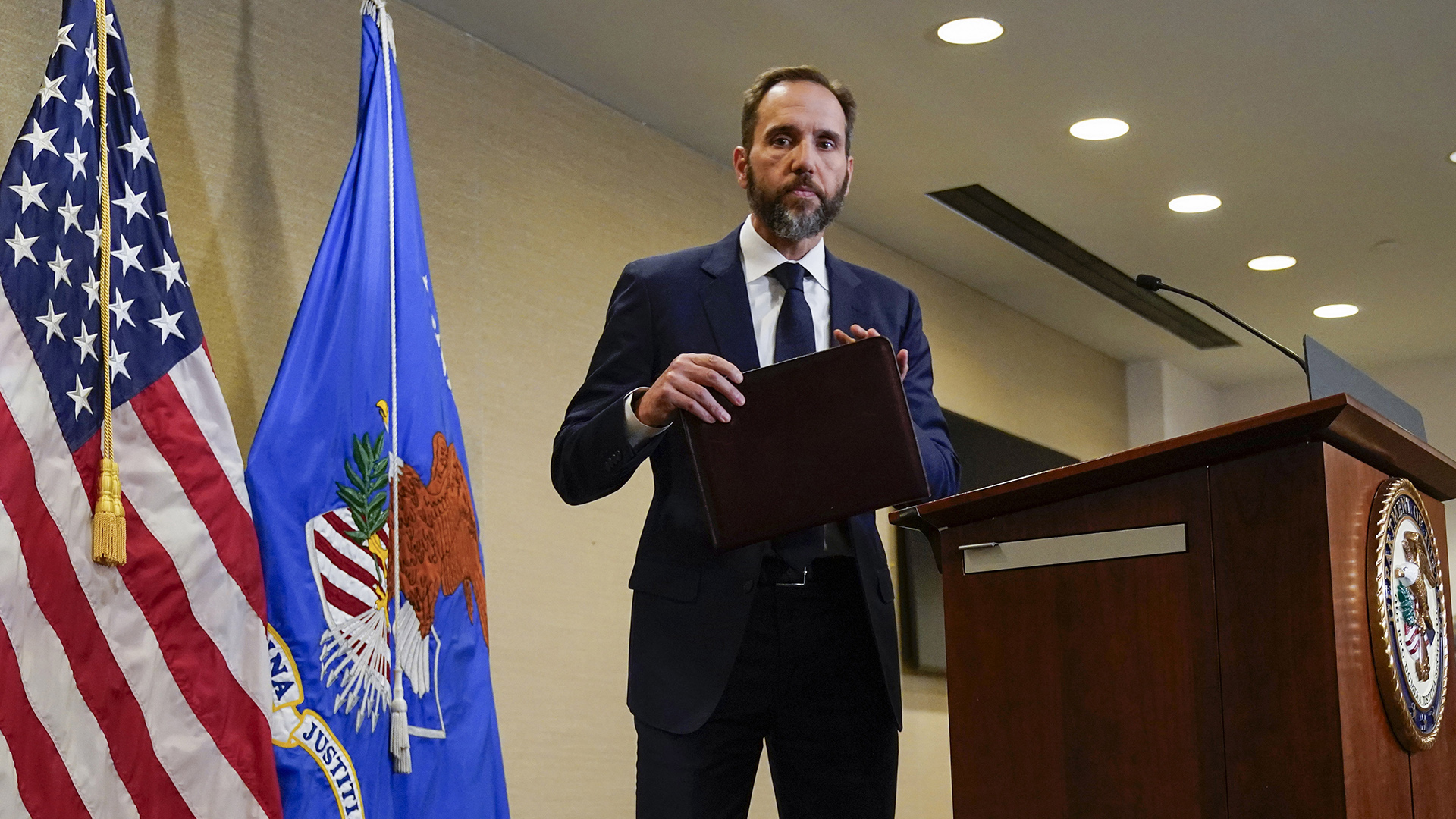 Jack Smith holds a legal brief folder in his hands while stepping away from a wood podium affixed with a microphone and the seal of the U.S. Department of Justice, in a room with recessed fluorescent lighting and the U.S. and Department of Justice flags.