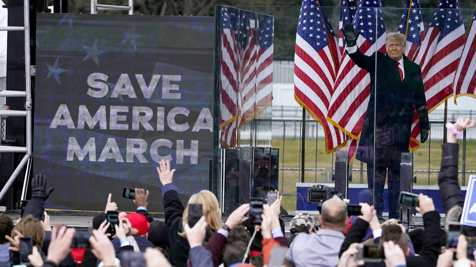 Donald Trump holds his right hand up in the air while standing on a stage and behind transparent barriers, next to an electronic screen showing the words Save America March and in front of a row of U.S. flags, with people facing the stage and holding their hands, cameras and cell phones in the air.