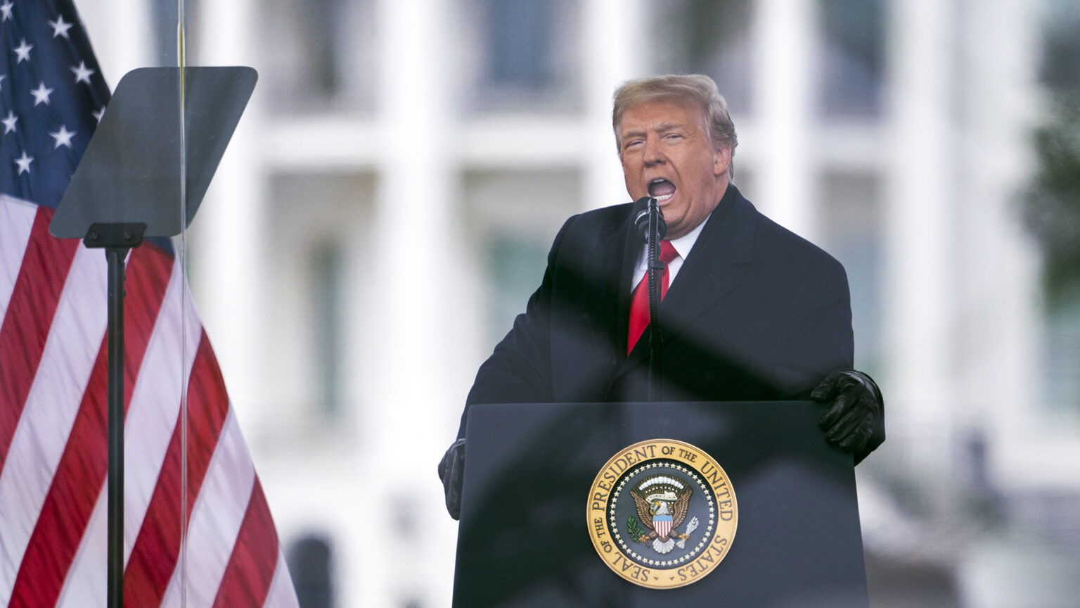 Donald Trump stands and speaks into a microphone mounted to a podium affixed with the Seal of the President of the United States while facing a teleprompter, with a U.S. flag and an out-of-focus White House in the background.